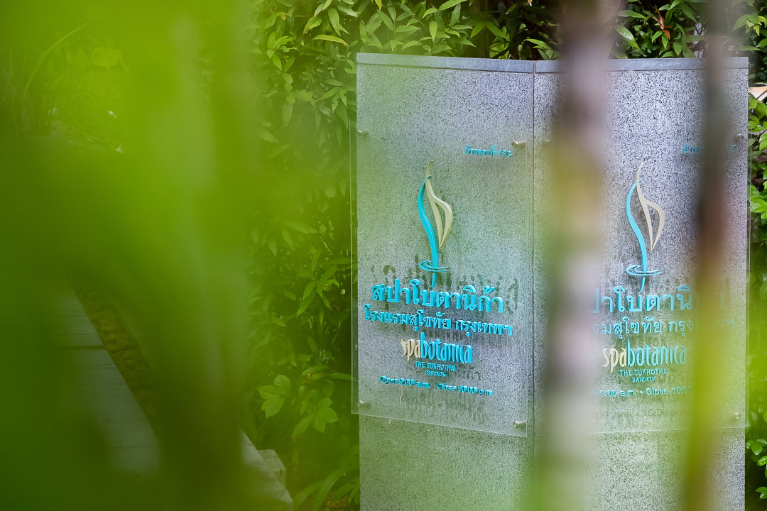 https://hungryformore.be/app/uploads/2024/12/sukhothai-bangkok-hotel-emblem-on-glass-behind-greenery.jpg