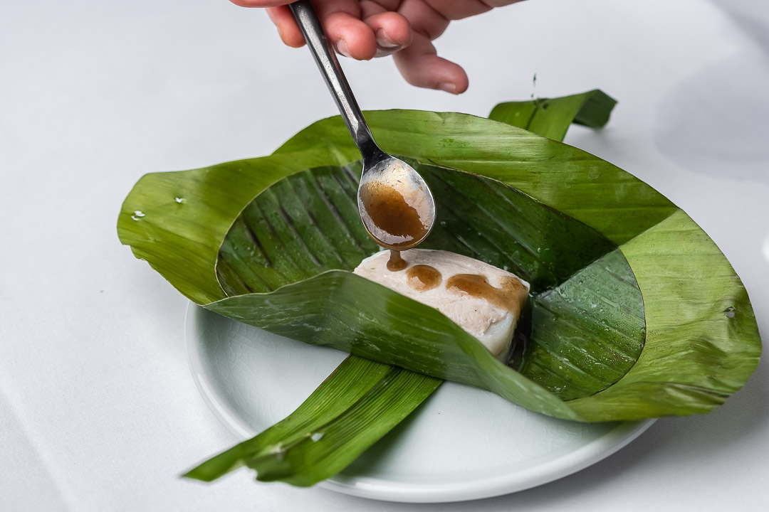 https://hungryformore.be/app/uploads/2024/12/sukhothai-bangkok-sauce-being-spooned-onto-a-banana-leaf-parcel.jpg